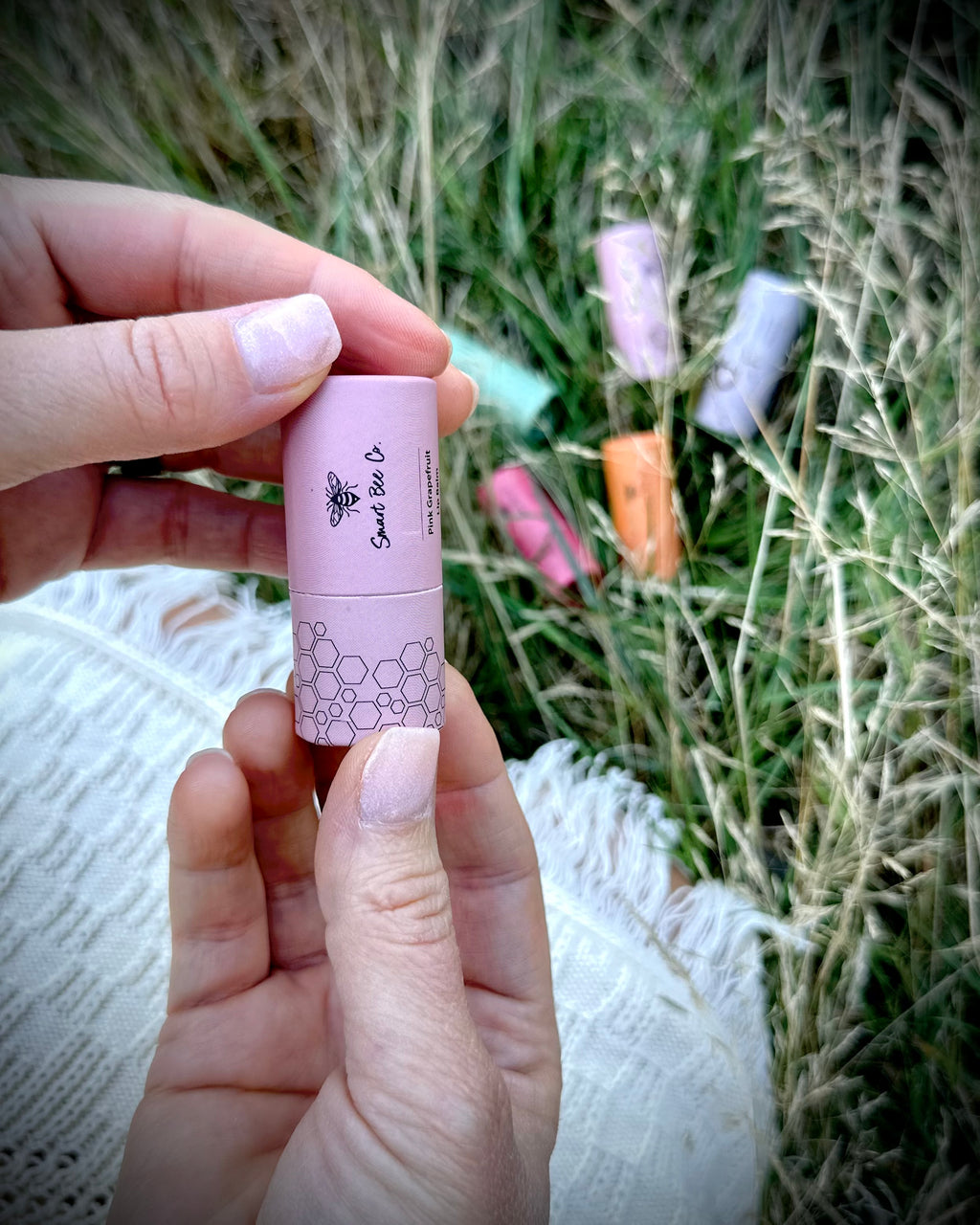 Hand holding a pink lip balm with blurred grass and other lip balms in the background