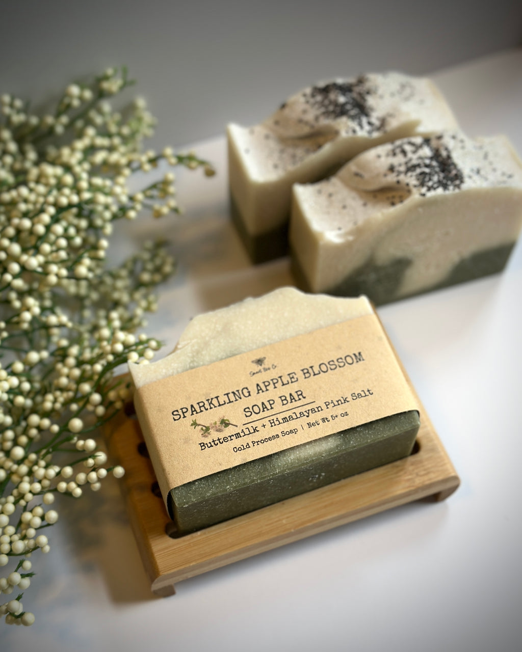 Bar of soap with a label on a wooden stand, surrounded by small white flowers.