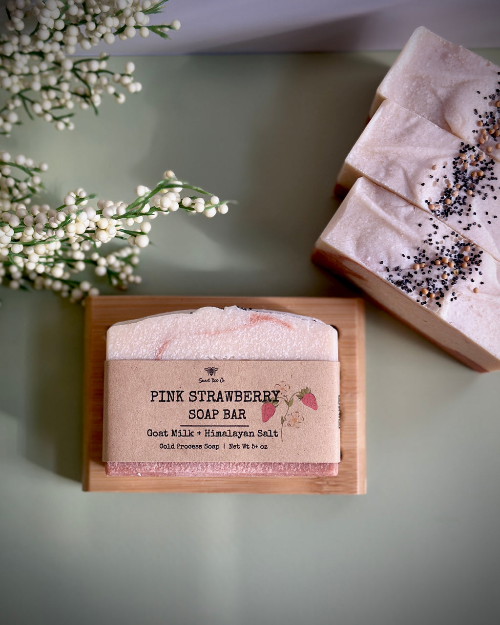Pink strawberry soap bar on a wooden block with a label, surrounded by small white flowers.
