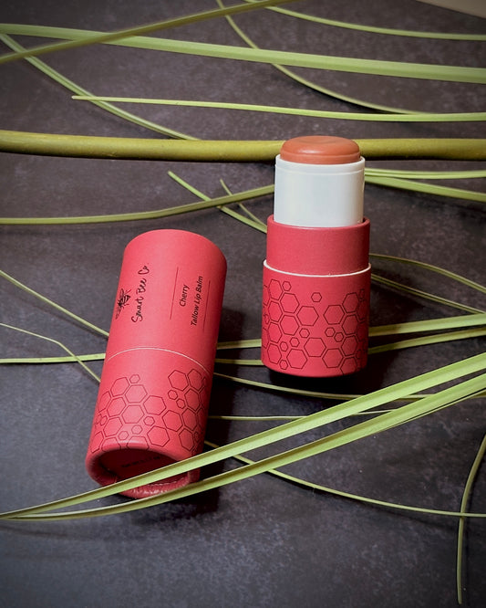 Two cherry tallow lip balm sticks with hexagonal patterns on a dark surface with green leaves.
