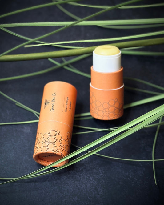 Two organic-looking skincare lip balm products on a dark background with green leaves.