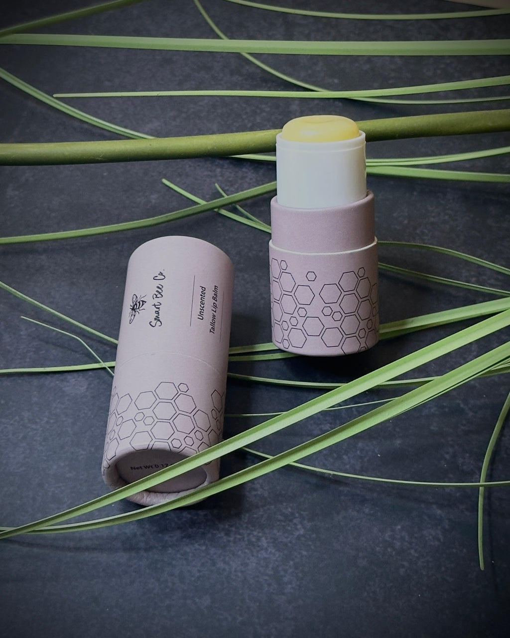 Two cylindrical lip balms products with honeycomb design on a dark surface with green leaves.