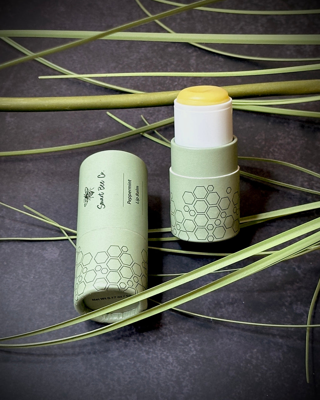 Two peppermint lip balm products with honeycomb design on a dark background with green leaves.
